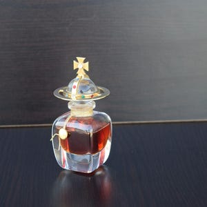 Op de afbeelding: Een heldere glazen fles met een gouden dop en een kruisontwerp. De fles is gevuld met een donkerbruine vloeistof.