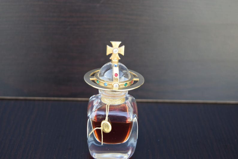 Op de afbeelding: Een doorzichtige glazen fles met een gouden bolvormige dop en een gouden kruis bovenop. De fles bevat een donkere vloeistof en heeft een gouden etiket.
