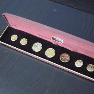 Op de afbeelding: Een rode fluwelen doos met een gouden label met de tekst "Vermeil" en acht gouden manchetknopen met verschillende initialen.