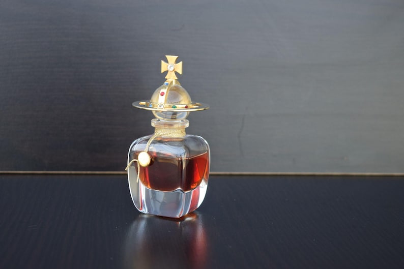 Op de afbeelding: Een doorzichtige glazen fles met een gouden kruis en een gouden kroon op de bovenkant. De fles is gevuld met een donkere vloeistof en staat op een zwart oppervlak.
