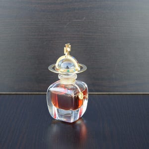 Op de afbeelding: Een doorzichtige glazen fles parfum met een gouden dop en een bolvormig ontwerp. De fles is gevuld met een donkerbruine vloeistof.