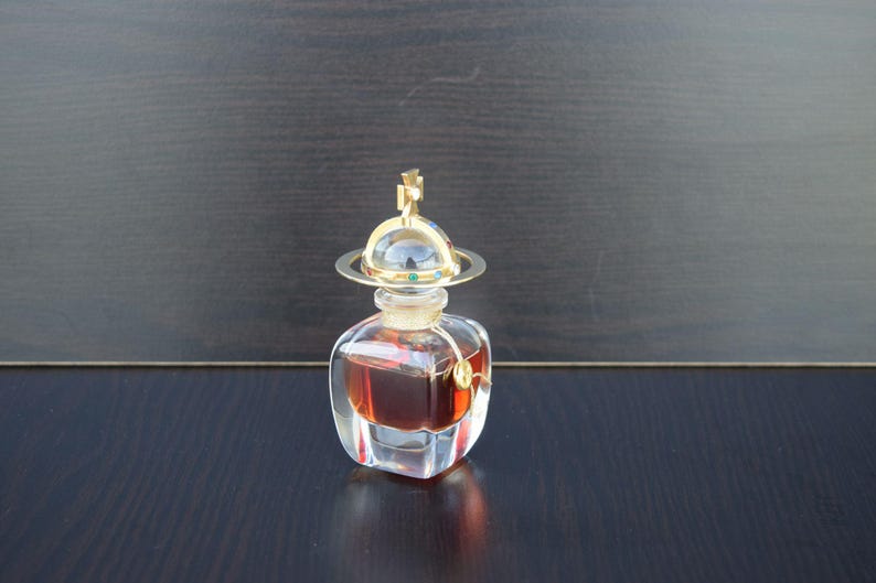 Op de afbeelding: Een doorzichtige glazen fles parfum met een gouden dop en een bolvormig ontwerp. De fles is gevuld met een donkerbruine vloeistof.
