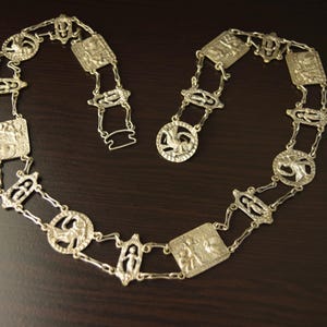 May include: Silver-coloured metal necklace with rectangular and circular decorative links. The necklace features detailed relief designs of figures and symbols. The clasp is open, and the necklace is displayed on a dark brown surface.