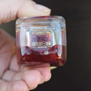 Op de afbeelding: Een klein, doorzichtig glazen flesje met een rode vloeistof erin. Het flesje heeft een gouden etiket met de tekst "0.67 FL OZ e 20 ML PARFUM".