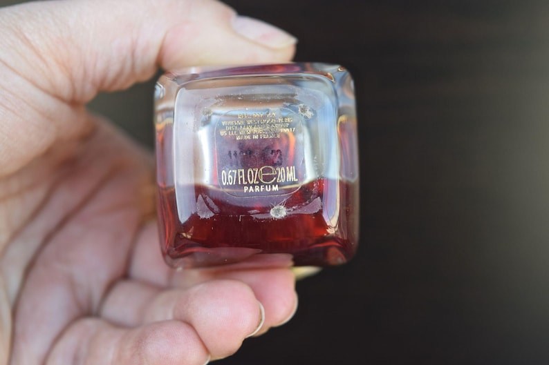 Op de afbeelding: Een klein, doorzichtig glazen flesje met een rode vloeistof erin. Het flesje heeft een gouden etiket met de tekst "0.67 FL OZ e 20 ML PARFUM".