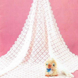 Puede incluir: Manta de bebé de ganchillo blanco con un delicado borde de encaje. Un pequeño juguete de peluche con forma de pollito está sentado sobre la manta.