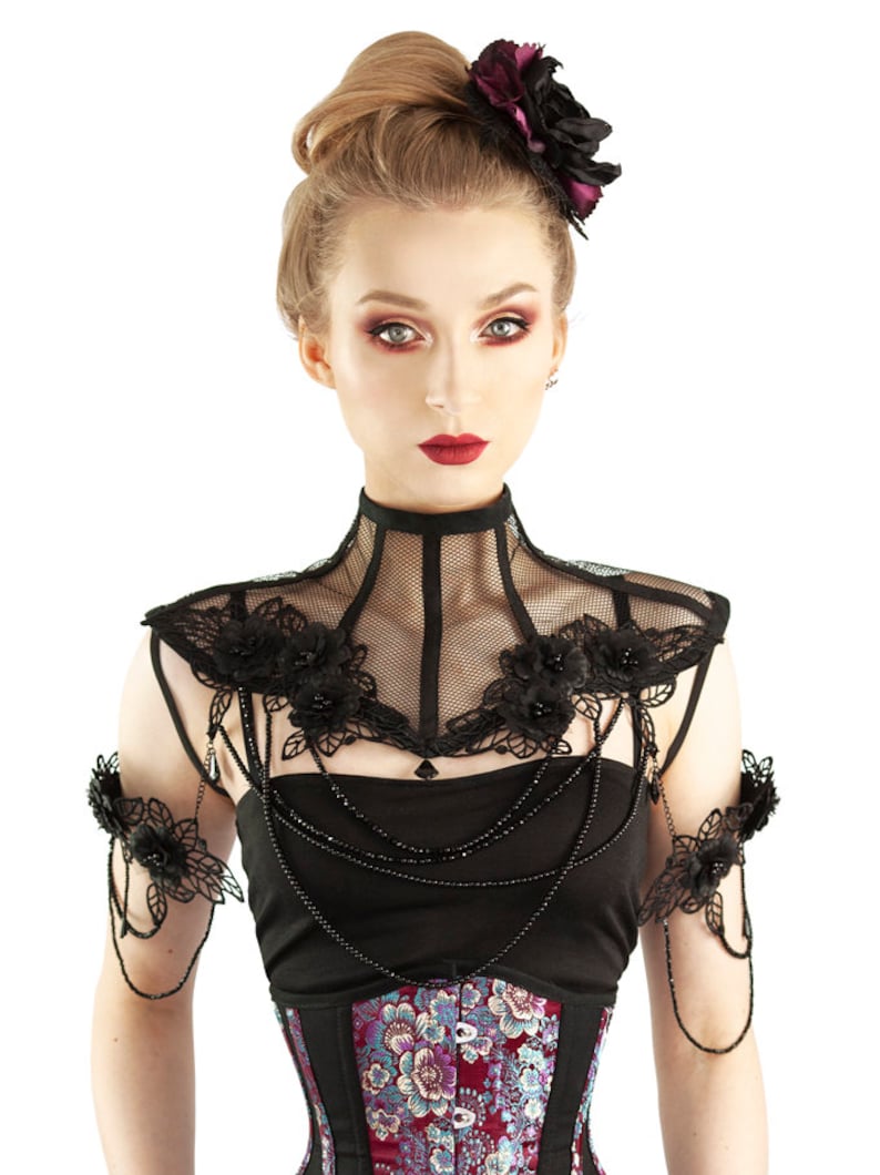 Puede incluir: Un collar de encaje negro con adornos florales y un par de pu&ntilde;os a juego. El collar tiene un dise&ntilde;o de malla y est&aacute; adornado con cuentas negras. La modelo lleva una blusa negra y un cors&eacute; con estampado floral.