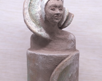 May include: A ceramic sculpture of a woman with a white and green abstract headpiece. The sculpture is brown and has a textured surface.