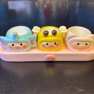 Può includere: Un set di tre piccoli contenitori in ceramica rotondi e cavi a forma di facce di animali, tra cui un elefante azzurro, un'ape gialla e un orso bianco, poggiati su un vassoio rettangolare rosa chiaro con un dettaglio a forma di muso di maiale.