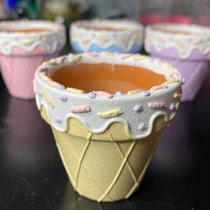 Peut inclure: Un ensemble de quatre petits pots en terre cuite peints pour ressembler à des cornets de glace. Chaque pot présente une base jaune texturée en gaufre, un bord blanc façon glaçage coulant et des vermicelles roses, jaunes et violets.