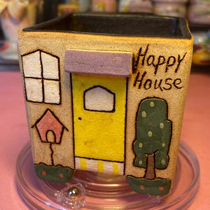 May include: A square ceramic planter with a beige textured finish featuring a yellow door, a purple awning, a window, a pink birdhouse, and a green tree. The text "Happy House" is written in black on the front. The interior is black.