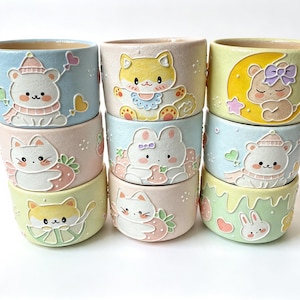 Può includere: Una collezione di otto piccoli vasi cilindrici in ceramica dai colori pastello, ognuno caratterizzato da illustrazioni in rilievo di animali in stile cartoon come orsi, gatti e conigli, arricchiti da motivi come fragole, lune e cuori.
