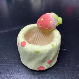 Può includere: Un piccolo contenitore cilindrico verde chiaro dalla consistenza opaca, decorato con punti rosa e gialli sparsi. Una fragola scolpita con gambo verde e semi gialli è fissata sul bordo superiore, sporgendo leggermente sopra l'apertura.