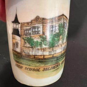 May include: A white ceramic tumbler with a detailed illustration of a school building and a church tower. The text "HIGH SCHOOL SOLDIERS GR" is printed below the illustration. The tumbler has a slightly flared shape.