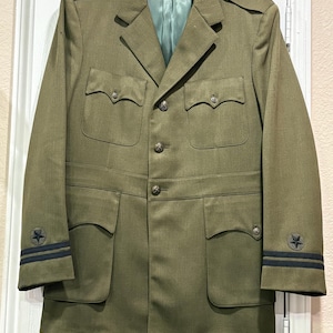 May include: Olive green military jacket with four front pockets, button closures, and a light green satin lining. The sleeves have black stripes and a star emblem. The jacket is hanging on a wooden hanger.