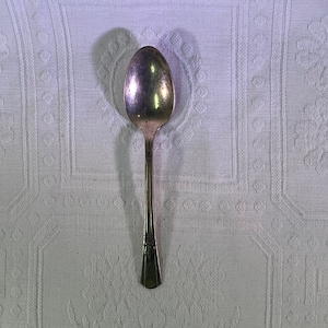 May include: A silver teaspoon with an ornate handle. The spoon is on a white tablecloth with a textured pattern.
