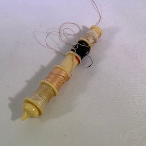 May include: A vintage white plastic spool holder with three spools of thread. The spools are wound with white, peach, and black thread. The thread is partially unwound.