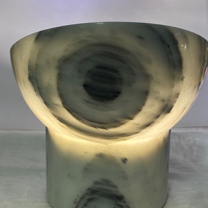 May include: A distinctive lamp featuring a marble design, with a cylindrical base and a bowl-shaped top. The lamp emits a warm light, accentuating the swirling grey and white patterns. It has a power cord.