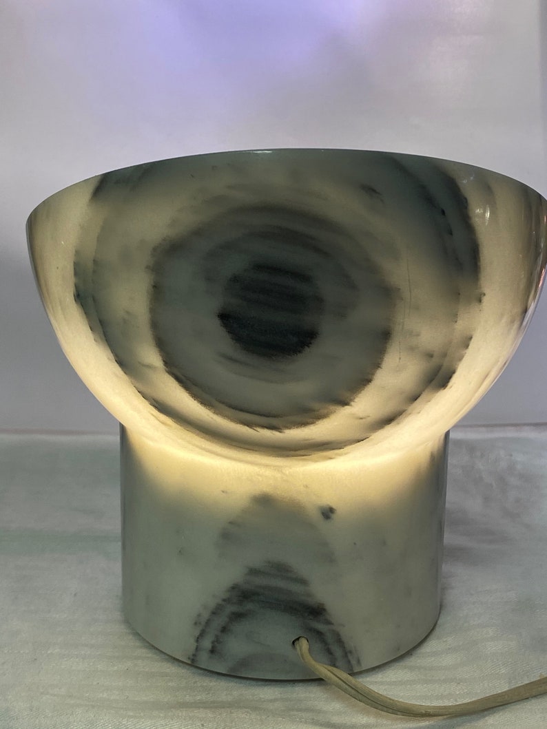 May include: A distinctive lamp featuring a marble design, with a cylindrical base and a bowl-shaped top. The lamp emits a warm light, accentuating the swirling grey and white patterns. It has a power cord.