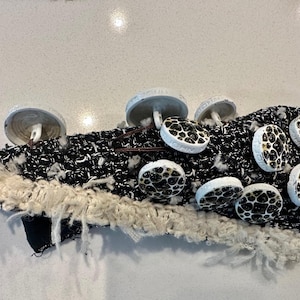 May include: A textile art piece featuring a black and white woven base with a cream-colored fringe. It is adorned with several mushroom-shaped embellishments with a white cap and a black and tan spotted pattern.