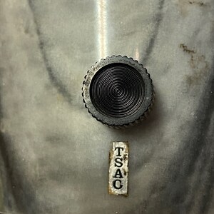 May include: Close-up of a metal object featuring a textured, circular knob and a label reading "TSAC". The object has a marbled, grey and white surface. The knob is black with concentric circles.