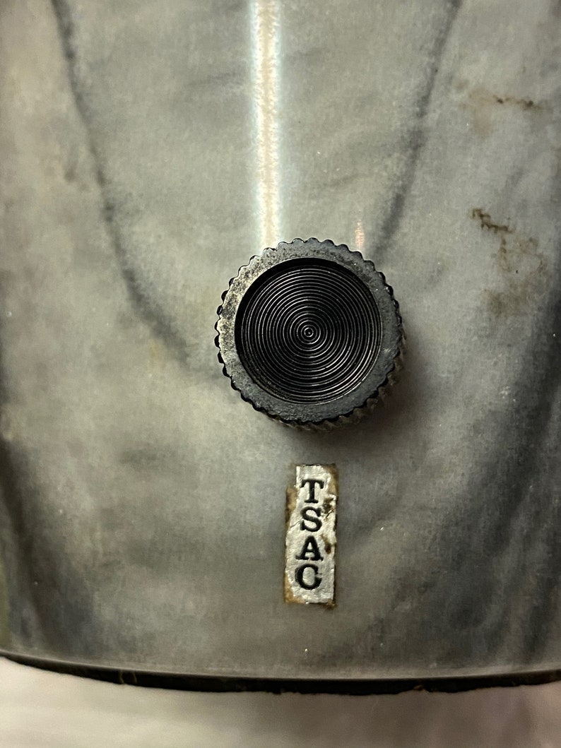 May include: Close-up of a metal object featuring a textured, circular knob and a label reading "TSAC". The object has a marbled, grey and white surface. The knob is black with concentric circles.