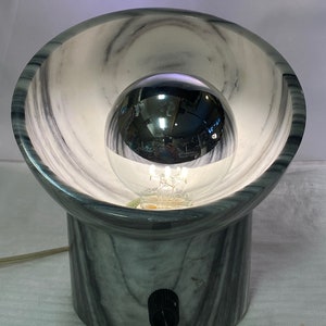 May include: A contemporary table lamp with a grey and white marble effect. The lamp has a spherical bulb within a bowl-shaped shade. A black switch is on the base. The lamp is plugged into an electrical outlet.