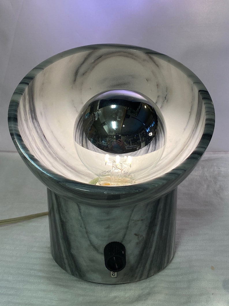 May include: A contemporary table lamp with a grey and white marble effect. The lamp has a spherical bulb within a bowl-shaped shade. A black switch is on the base. The lamp is plugged into an electrical outlet.