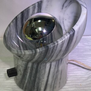 May include: A distinctive lamp with a marble base and a chrome sphere. The lamp has a grey and white marble design with a spherical chrome accent. It features a black knob and a power cord. The lamp's design is modern and artistic.