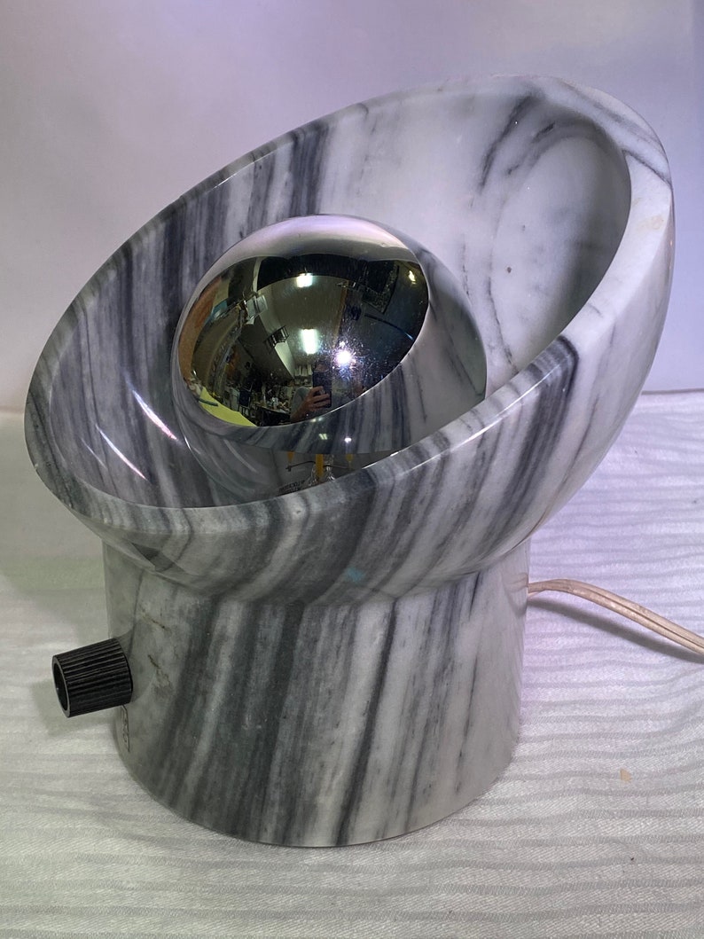 May include: A distinctive lamp with a marble base and a chrome sphere. The lamp has a grey and white marble design with a spherical chrome accent. It features a black knob and a power cord. The lamp's design is modern and artistic.