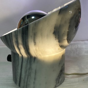 May include: A contemporary table lamp with a marble base and a curved, bowl-shaped top. The lamp has a reflective, spherical light source and a black on/off switch. The design incorporates a mix of white and grey marble.