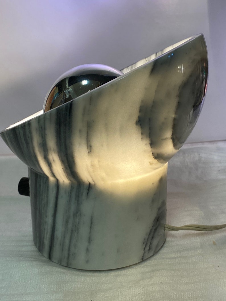 May include: A contemporary table lamp with a marble base and a curved, bowl-shaped top. The lamp has a reflective, spherical light source and a black on/off switch. The design incorporates a mix of white and grey marble.