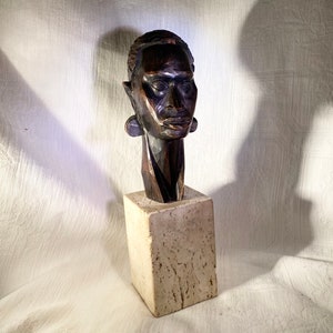 May include: A dark brown wooden sculpture of a human head with large earrings, mounted on a square, light-coloured stone base. The sculpture has a smooth, polished finish and is set against a white background.