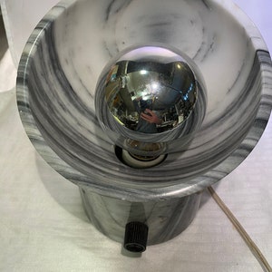 May include: A marble-effect table lamp featuring a spherical chrome bulb. The lamp has a bowl-shaped shade and a cylindrical base with a black on/off switch. The design incorporates grey and white veining.