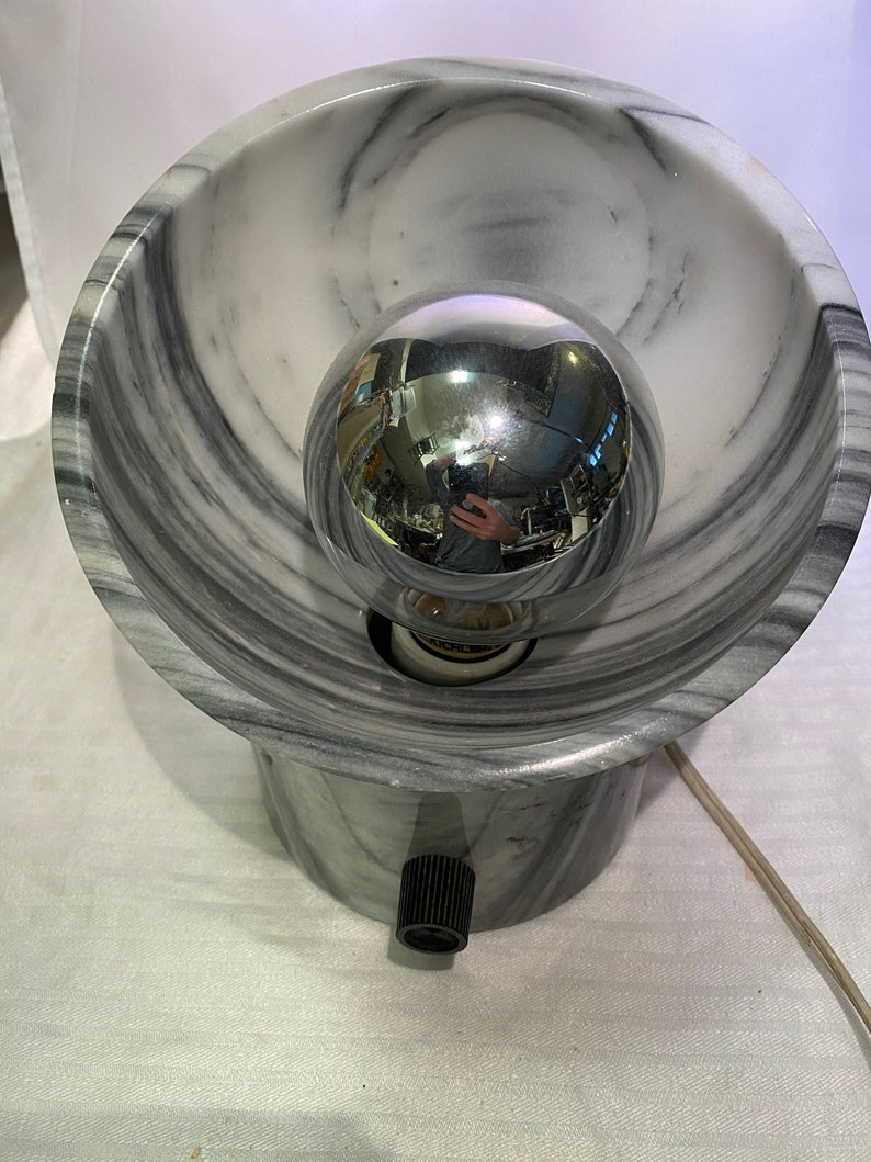 May include: A marble-effect table lamp featuring a spherical chrome bulb. The lamp has a bowl-shaped shade and a cylindrical base with a black on/off switch. The design incorporates grey and white veining.