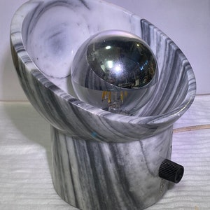 May include: A contemporary table lamp featuring a chrome spherical bulb encased in a grey and white marble oval shade. The lamp has a cylindrical base and a black knob for adjusting the light. The lamp is plugged into an electrical outlet.