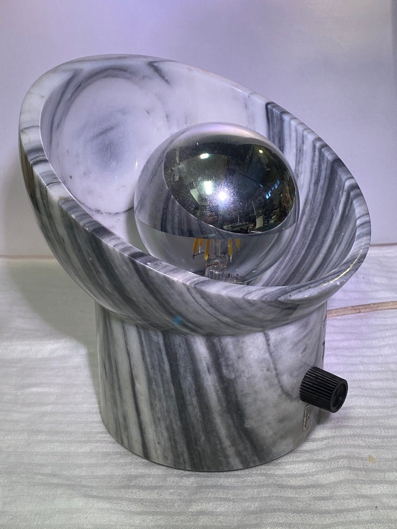 May include: A contemporary table lamp featuring a chrome spherical bulb encased in a grey and white marble oval shade. The lamp has a cylindrical base and a black knob for adjusting the light. The lamp is plugged into an electrical outlet.