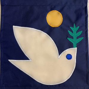 Puede incluir: Una bandera de tela azul marino con una paloma blanca que lleva una rama de olivo verde. La paloma tiene un ojo azul y está bordeada de blanco. Un círculo amarillo se encuentra en la esquina superior derecha de la bandera.