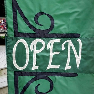 May include: Green fabric banner with black and white lettering that reads "OPEN". The banner has two black decorative swirls on either side of the word "OPEN".