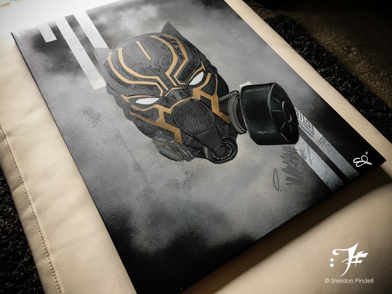 Puede incluir: Obra de arte en aerosol negro y dorado de una m&aacute;scara de Black Panther con una m&aacute;scara de gas. La obra de arte est&aacute; en un lienzo y tiene un fondo blanco con l&iacute;neas negras.