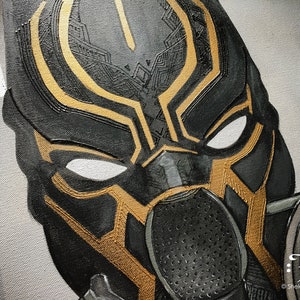 Puede incluir: Primer plano de una m&aacute;scara de superh&eacute;roe negra y dorada con un dise&ntilde;o detallado. La m&aacute;scara tiene un &aacute;rea de ojos blanca y un dise&ntilde;o estampado negro y dorado.