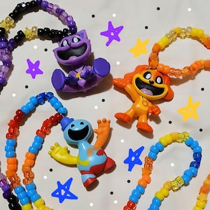May include: Three colorful plastic character figurines attached to beaded necklaces. One purple cat-like figure, one orange dog-like figure, and one blue and red clown-like figure, each with a wide smile, are strung on strands of multi-colored plastic pony beads.