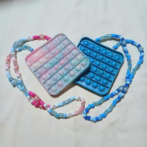 May include: Two square silicone sensory fidget toys with rows of circular bubbles. One toy features a pastel tie-dye pattern of pink, blue, and white, while the other is solid blue. Each toy is attached to a long, colorful beaded necklace strap.