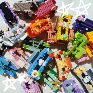 Könnte beinhalten: Eine Sammlung von Haarklammern aus Kunststoff in verschiedenen Farben wie Blau, Pink, Grün, Gelb und Lila. Jede Klammer ist mit Miniatur-Harzfiguren verziert, darunter Zeichentrickfiguren, Blumen, Sterne, Früchte und Süßigkeiten.