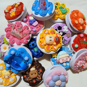 May include: A collection of round phone grips featuring decorative tops with colorful whipped cream-like textures, miniature character figurines, rhinestones, and small charms in various themes including animals, fruits, and fantasy motifs.