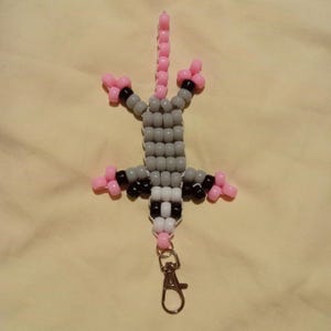 Könnte beinhalten: Ein Schlüsselanhänger mit einem Opossum aus Perlen in Grau, Schwarz, Weiß und Rosa. Das Opossum besteht aus kleinen Perlen und hat einen rosa Schwanz und Füße. Es hat einen silberfarbenen Verschluss zum Befestigen an Schlüsseln oder einer Tasche.