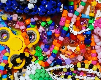 Può includere: Primo piano di una collezione di gioielli e accessori colorati con perline. L'immagine presenta braccialetti, collane e altri oggetti in una varietà di colori vivaci, tra cui rosa, blu, giallo e verde. È visibile anche un fidget spinner giallo.