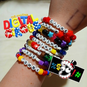 Puede incluir: Una colección de pulseras de cuentas coloridas que deletrean nombres y palabras, incluyendo "CAINE", "ANGE", "ORBSMA", "GLOINK", "INGE" y "POMNI". Las pulseras presentan cuentas de varios colores y acentos en forma de estrella. La imagen también incluye el texto "THE AMAZING DIGITAL CIRCUS" y un gráfico con símbolos.