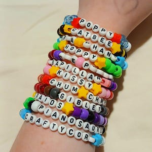 May include: A collection of colorful beaded bracelets spelling out various words like "POPPY", "CATBEE", and "CRAFTYCOR". The bracelets feature white letter beads, star-shaped beads, and a variety of other colorful beads. The bracelets are stacked on a wrist.