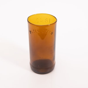 May include: A brown glass drinking glass with the text "PLZEN CZECH" etched on the side. The top of the glass is yellow.